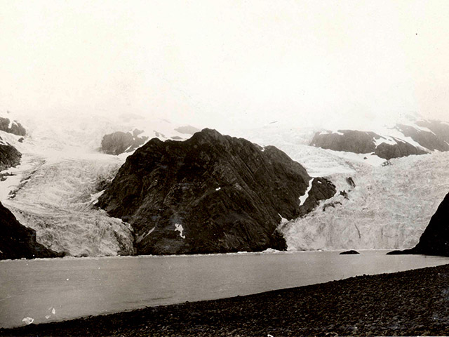 A 1909 black and white photo of a dark mountain peak surrounded by a stark white glacier on each side that flows into a body of water. As the slider handle moves to the right, the photo turns into a color photo from 2005 of the same mountain peak. Valleys are on either side with much less glacier ice.