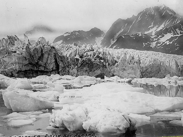 A 1909 black and white photo of dark mountains with a forested hillside on the right, a large glacier on the left, and a lake in the foreground. As the slider handle moves to the right, the photo turns into a color photo from 2005 of the same location, but the glacier is entirely gone in this view, and green vegetation covers over half of the lake.