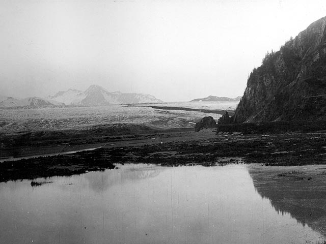 A 1909 black and white photo of snow-topped mountains, a glacier in front of a rocky flat and a log.  As the slider handle moves to the right, the photo turns into a color photo from 2005 of the same mountains with very little snow on them. The glacier is not visible; trees and vegetation are in its place. The log is still there.