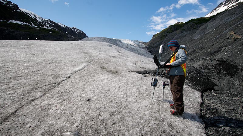 A park staff member stands with equipment at the edge of a glacier entering data on a laptop computer.