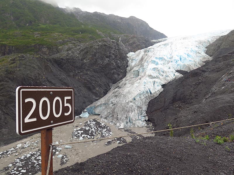 Exit Glacier terminus and outwash plain are nestled in the surrounding hilly topography. A sign marked with the year 2005 marks the location of the glacier's terminus in 2005.