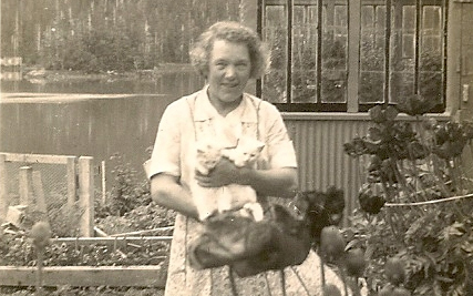 Josephine Sather stands among tall flowers in her garden holding two white kittens.