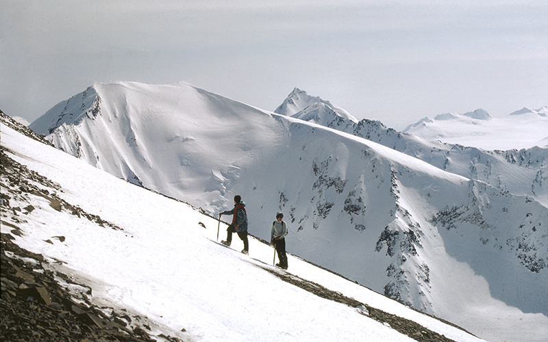Two mountaineers climb up a snowy slope in front of mountains surrounding the Harding Icefield in 1968.