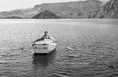 A man in a hat rows a boat on a lake with mountains in the background.