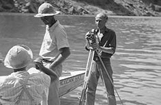 Man stands on small boat with a video camera on a tripod, while two people stand nearby.