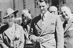 Woman in a hat with gloves and purse stands next to man in a suit.