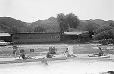 Four women lounge by pool in front of building while two people swim.