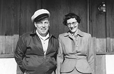 Man with a hat poses with a woman in a suit and glasses.