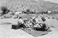 Four people picnicking on a blanket in foreground with small mountain and several cars in the background.