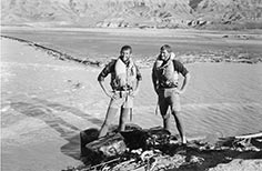 Two men stand beside several bags with hands on hips wearing life vests next to a lake with mountains behind them.