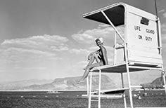 Woman in swimsuit sits on life guard stand overlooking a lake.