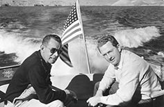 Two men sit in a boat on a lake with an American flag attached.
