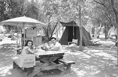 Man and woman sit on picnic table in a campsite with a tent and awning.