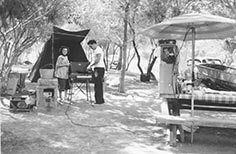 Man and woman stand by barbeque grill in a campsite with a tent, awning, and picnic table.