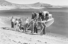 People stand on and in front of a boat tied to shore, one man looking through a video camera.