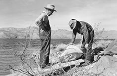 A man standing with hands on hips looking down towards another man with hands on a large rock with petroglyphs.