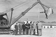 Several men in suits stand in a group next to a bulldozer.