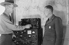 Two men in uniform stand in front of a communication machine with a map on the wall behind them.