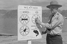 Man in uniform stands next to weather sign with dials on it.