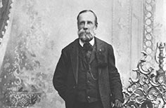 Man with a beard stands posing next to an ornate chair.