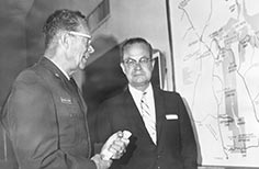 Two men stand by framed map on a wall.