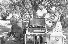 Four people sit and stand around a portable stovetop on a picnic table.