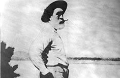 Bearded man stands with hand in pocket and a hat smoking a cigarette.