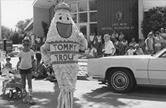 A crowd of people stand and sit in front of a building with a person in a fish shaped mascot costume out front.