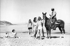 Man in uniform rides a horse on a beach while two women in swimsuits look over from sun bathing and more women pet the horse.