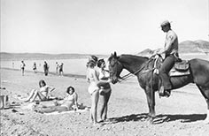 Man in uniform rides a horse on a beach while two women in swimsuits look over from sun bathing and two women pet the horse.