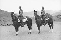 Two men in uniform pose on horses.