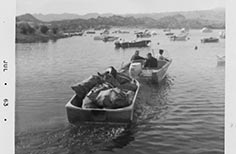 People in a boat tow another boat full of bags on a lake with other boats in the distance.