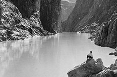 Landscape view of a creek surrounded by towering rocks while a man in a hat sits on a smaller rock formation on the bottom left, facing the view.