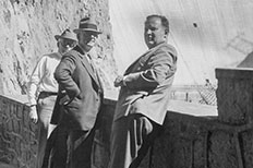 Three older men dressed nicely and posing for the camera along a wall in front of the Boulder Dam.