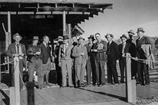 Thirteen men in suits and hats standing beside each other as they pose on a wooden dock.