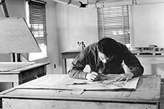 Dark-haired gentleman keeps his head down as he traces an outline of a design on a rectangular wooden table.