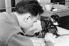 Side-profile of a man with his head down as he uses both hands on a small black machine to cut out a section in the construction of a relief model.