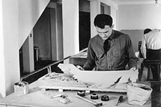 Dark-haired man with his head down as he places a piece of finished contour in the relief model, with tool-like objects scattered across the rest of the table.