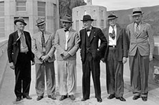 Six older men stand side by side in suits and panama hats as they pose in front of two cylinder shaped buildings with windows.
