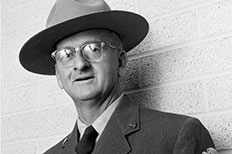 Older gentleman wearing glasses, hat, and suit with a NPS badge on the right sleeve with a textured brick wall behind him.