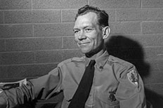 Older gentleman smiling while wearing a ranger uniform with a pin on the pocket and NPS badge on the right sleeve, with his left arm resting on a table next to him in front of brick wall.