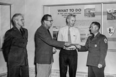 Four gentleman standing side by side as one hands over an award to a man on the end, with a poster board-like image of ‘What to do in the Lake Mead National Recreation Area’ displayed behind them.