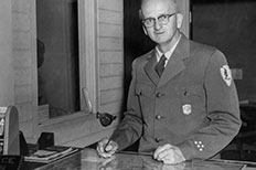 Gentleman dressed in NPS ranger uniform with a badge on the pocket and NPS embroidered patch on the right sleeve, hands rest on a table, a pen in his left hand.