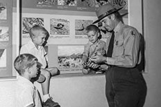 Two little boys sit on the ledge of display full of wildlife and landscape photos, the third little boy stands with a ranger dressed in uniform as he holds a snake to show them.