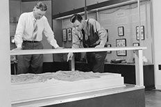 Two gentleman looking down at a relief model in the NPS museum, one having his hands on top of the model while the other uses his left hand to point at it.