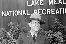 Gentlemen smiles in park ranger uniform as he stands in front of plants and wooden textured wall, only capturing the words ‘Lake Mead National Recreation’ in the background.