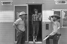 A trailer house with a woman standing in the doorway as two gentlemen in differing uniforms stand outside on either side of her having a conversation.