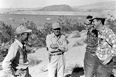 Four gentlemen facing each other as they have conversation with a background of desert, surrounding mountains, and a body of water with boats scattered across.