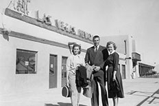 Gentleman in a suit with two women on each side of him as they all hold luggage in front of a building with a sign at the top that reads Las Vegas Nevada, with a cowboy and horse icon.