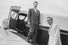 Two gentleman in suits standing on the wing of an airplane near cockpit door, one gentleman higher above the other as his hand rests on the door handle.